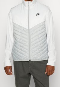 Nike är en ärmlös jacka med vita och ljusgrå färgblock, vadderad yta, hög krage och framfickdragkedja. Svart logotypdetalj finns.