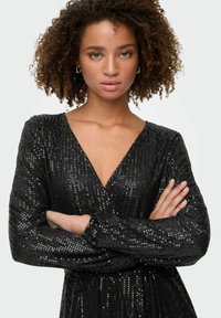 Crna haljina s nijansama, dubokim V-izrezom, dugim rukavima i uskim strukom. Tekstura je glatka s reflektirajućom površinom od šljokica.