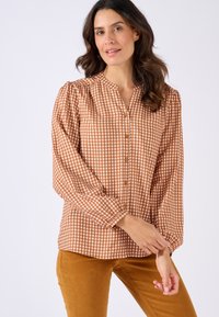 Blusa a scacchi marrone e crema con chiusura a bottoni, maniche a sbuffo e collo arricciato. Abbinata a pantaloni color senape. Tessuto dalla texture liscia.