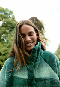 Veste en polaire verte avec un col haut et une finition texturée, présentant des nuances de vert variées et un détail de bouton au niveau du col.