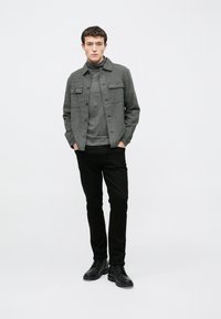 Jachetă gri cu model, cu două buzunare frontale, purtată peste un pulover gri cu guler înalt, asortată cu pantaloni negri mulați și bocanci negri cu șireturi.