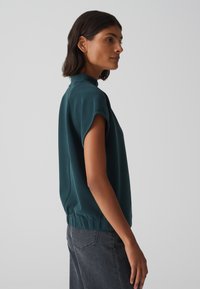 Haut teal à manches courtes avec un col haut, taille élastique, texture lisse et coupe décontractée, associé à un jean gris foncé.
