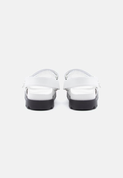 Λευκά σανδάλια με μαύρες χοντρές σόλες και ρυθμιζόμενους πίσω ιμάντες, εμφανιζόμενα από πίσω σε απλό λευκό φόντο.