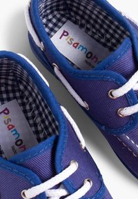 Zapatillas de lona moradas con cordones blancos, con un forro a cuadros en blanco y negro. Etiqueta de la marca visible en el interior. Los detalles incluyen ojales de metal.