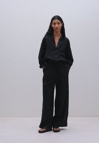 Fekete gombos ing hosszú ujjakkal, széles szárú fekete nadrággal párosítva. Simább anyag, laza illeszkedéssel. Egyszerű dizájn, minimális részletezéssel.