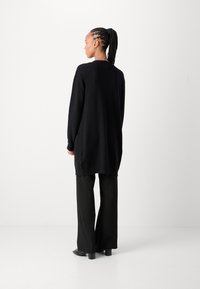 Long gilet noir avec une coupe décontractée, présentant des poignets côtelés et un dos légèrement plus long, associée avec un pantalon large noir et des chaussures à talons.