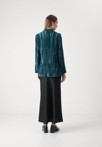 Teal velvet blazer with vertical stripes, worn over a long black skirt. The blazer features a notched collar and structured silhouette.