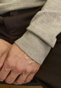 Jasnoszary, prążkowany mankiet swetra o miękkiej fakturze, nałożony na ciemnobrązowy materiał. Widoczna dłoń oparta na materiale poniżej.