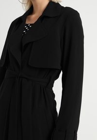 Kobieta w czarnym trenczu z paskiem noszącym czarno-białą bluzkę w kropki, pokazana od ramion do bioder na jednolitym tle.