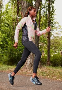 Completo sportivo per la corsa con una giacca a fantasia rosa e nera, leggings neri e scarpe da corsa grigie con accenti blu, ambientato in un'area boschiva.