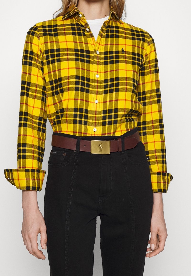 Personne portant une chemise à carreaux jaune et noire boutonnée, rentrée dans un pantalon noir avec une ceinture en cuir marron et une boucle dorée.