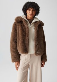 Bruine nep bontjas met een capuchon, met een ritssluiting aan de voorkant en bruine knopen. Draag zich over een crèmekleurig gebreid topje en beige hooggesneden broek.