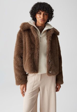 Braune Kunstfelljacke mit Kapuze, mit einem Reißverschluss vorne und braunen Knöpfen. Getragen über einem cremefarbenen Strickoberteil und beigen hoch taillierten Hosen.