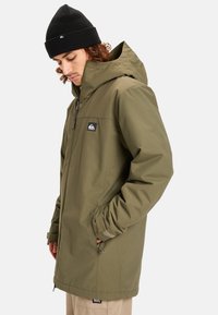 Olijfgroene, waterdichte jas met capuchon, ritssluiting aan de voorkant en logo patch. Voorzien van zijzakken en elastische manchetten. Draagt een zwarte beanie.