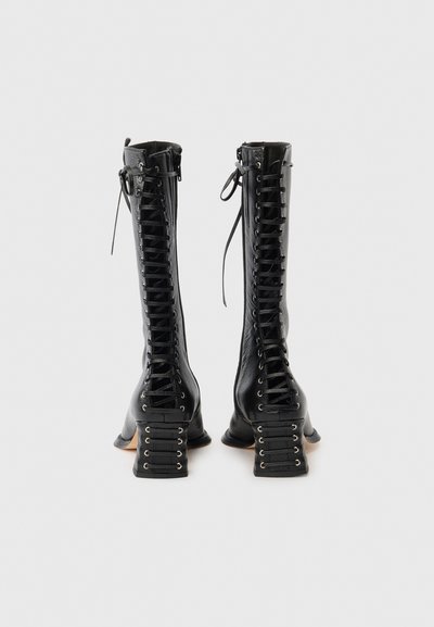 Μαύρες δερμάτινες μπότες μέχρι το γόνατο με περίπλοκο μπροστινό κορδόνι, χοντρά τακούνια τύπου block και διακοσμητικές λεπτομέρειες με κορδόνια στις πλευρές των τακουνιών.