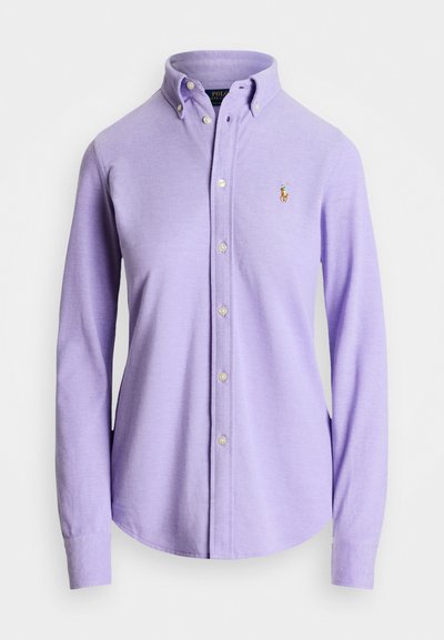 Camisa de manga larga de color púrpura claro con cuello abotonado, que presenta un pequeño logo bordado en el pecho y botones similares a perlas en la parte delantera.