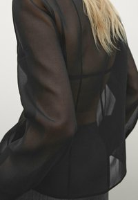 Musta läpinäkyvä paita, jossa on pitkät puffihihat ja istuva vyötärö. Ominaisuutena läpinäkyvä selkä sekä jämäkkä muotoiluelementti, jotka korostavat siluettia.