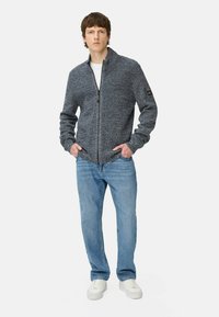 Grijze zip-up jack met een hoge kraag, gestructureerde gebreide stof en lange mouwen, gecombineerd met lichtblauwe jeans en witte sneakers.