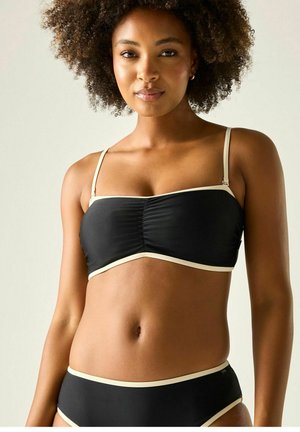 Frau mit lockigem Haar trägt einen schwarzen Bikini mit beigen Rändern, steht vor einem einfachen hellen Hintergrund und schaut in die Kamera.