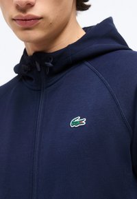 Νεαρός φοράει μπλε ναυτικό φούτερ με φερμουάρ, με πράσινο λογότυπο κροκοδείλου στο στήθος και κορδόνια στο κουκούλα.