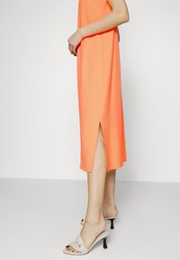 Světle oranžové bezramínkové šaty se bočními rozparky, vyrobené z jemné tkaniny. Nosené s béžovými sandály na klínku se uzlovým designem.