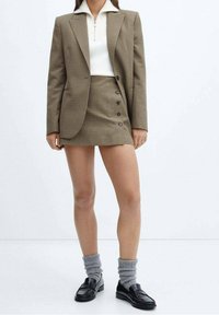 Blazer à carreaux marron avec des revers nets, associé à une mini-jupe assortie à boutons, un pull blanc à fermeture éclair, des chaussettes grises et des mocassins noirs.