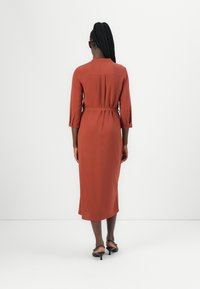 Robe midi couleur rouille avec des manches trois-quarts, une taille cintrée et un détail de pli au dos, fabriquée en tissu lisse.