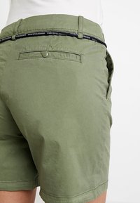 Olivově zelené šortky vyrobené z lehké látky. Mají zadní kapsu na knoflík a kontrastní pásek s logem.