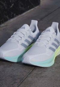 Paire de chaussures de course blanches avec des bandes grises et des semelles bleu-vert clair posées sur un sol en béton.
