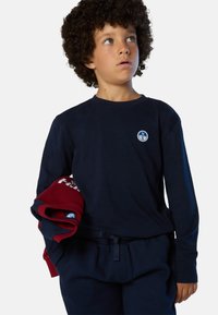 Camisa de manga larga azul marino con cuello redondo y parche de logo azul, combinada con pantalones jogger oscuros. Niño sosteniendo sudaderas roja y azul marino, con texto blanco.