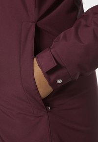 La chaqueta ligera burdeos presenta una textura suave, detalles de puños a presión y una apertura de bolsillo lateral, destacando un diseño elegante y minimalista.