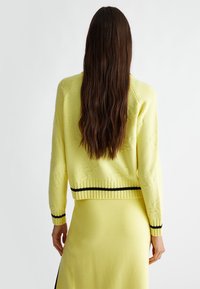 Maglione giallo lavorato a maglia con polsini e orlo a costine, caratterizzato da un design testurizzato e strisce nere lungo il bordo inferiore.