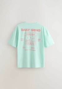 Lyseblå t-shirt med rød grafik af et kaffehus, teksten "Daily Grind," menupunkter og caféstole på bagsiden.