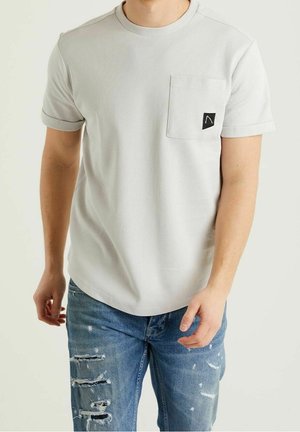 Homme portant un T-shirt texturé gris clair à manches courtes avec poche poitrine et un jean bleu déchiré, debout devant un fond uni.