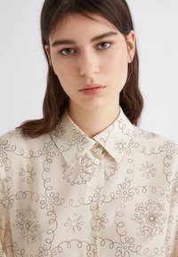 Blusa beige chiara con un motivo floreale sottile in marrone scuro, caratterizzata da un colletto a punta e chiusura con bottoni sul davanti.