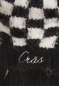 Crno-bijela karirana pletenina s pahuljastom teksturom, sa riječju "Crās" izvezanom bijelom niti.