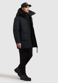 Geacă puffer albastră marină cu glugă, zippere frontal și logo pe piept. Purtați peste o ținută neagră și asortată cu cizme negre cu șireturi.