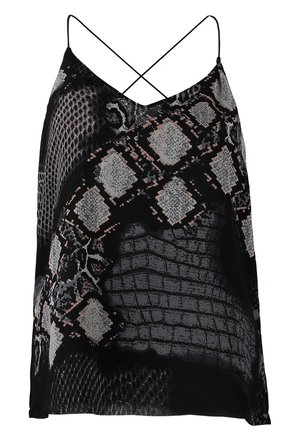 Kobieta w czarnym i szarym topie w wzór skóry węża, ciemnych dopasowanych dżinsach, beżowych szpilkach, trzymająca beżową kopertówkę.