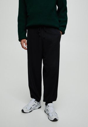 Persoon draagt zwarte broek met trekkoord, witte en zwarte sneakers en een donkergroene gebreide trui, staand met één hand in de zak.