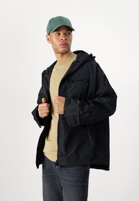 Svart vanntett jakke med hette, med glidelåslommer og justerbare ermer, paret med en beige skjorte og jeans, under en grønn caps.