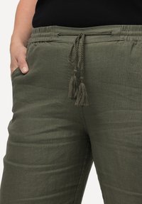 Pantalons vert olive, amples, avec une taille à cordon de serrage et des accents en pompon. Fabriqués en tissu texturé, avec des poches latérales.