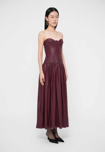 Bordové kožené šaty bez ramienok s priliehavým živôtikom a padajúcou, nabrúsenou sukňou, doplnené o čierne lodičky so špicatou špičkou.