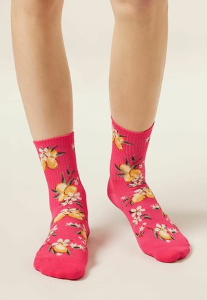 Jambes portant des chaussettes rose vif décorées de citrons jaunes et de fleurs blanches, posées sur un fond clair.