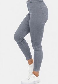 Primer plano de una persona que lleva jeans pitillo ajustados grises y zapatillas blancas, de pie contra un fondo blanco liso.