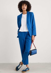 Niebieska marynarka i spodnie wykonane z teksturowanej tkaniny, w połączeniu z białą bluzką i granatowymi trampkami. Mała torba na ramię z czerwoną rączką.