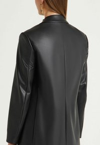 Blazer in finta pelle nera, design sartoriale con una texture liscia, colletto a lancia e spalle strutturate, che mette in risalto una silhouette elegante.