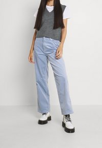 Maglione grigio lavorato a maglia senza maniche sopra una maglietta bianca, abbinato a pantaloni a gamba larga in velluto a coste blu chiaro e stivali bianchi con lacci e tacco alto.