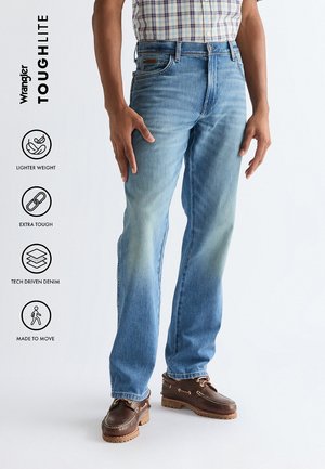 Man draagt lichtblauwe spijkerbroek en bruine schoenen, staand tegen een witte achtergrond met Wrangler ToughLite-functies vermeld.