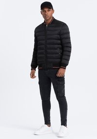 Giacca a vento nera con polsini e colletto a costine, con accenti rossi. Abbinata a pantaloni cargo neri slim fit e sneakers bianche.