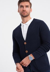Cárdigan de punto azul marino con escote en V, botones de madera y bolsillos delanteros, llevado sobre una camisa blanca con cuello. Reloj plateado en la muñeca.